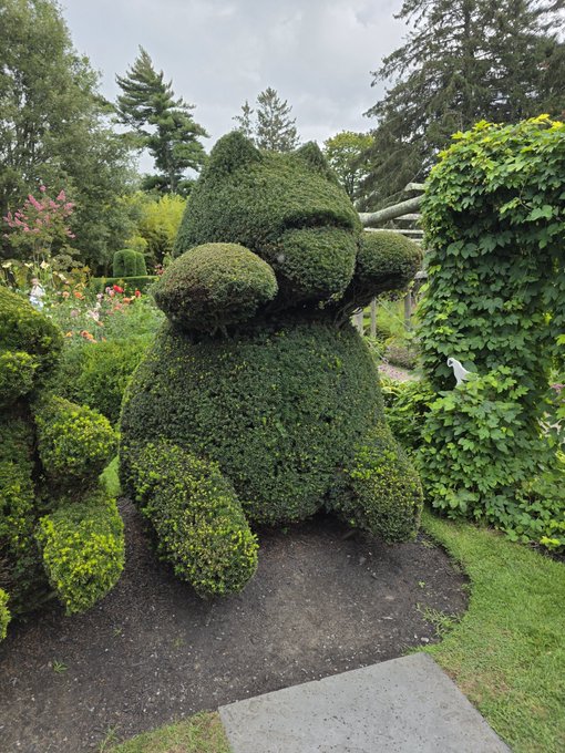 A large topiary sculpture of a cartoonish green creature sits in a garden surrounded by trees and foliage.