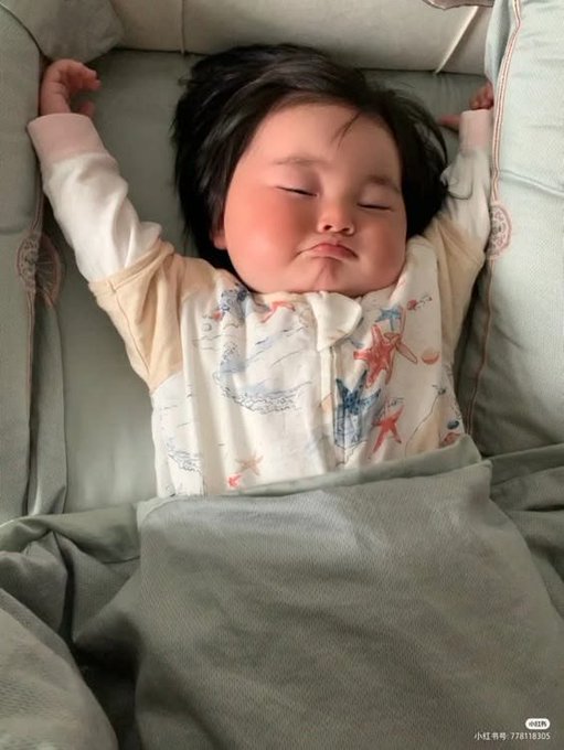 A sleeping baby with rosy cheeks and dark hair, wearing a patterned white onesie with sea creatures, lies with arms stretched up on a gray pillow.