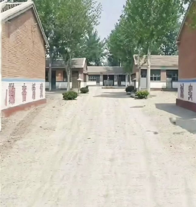 A dirt road leading into what appears to be a rural Chinese compound or village, with brick buildings on either side and Chinese characters painted on the walls.