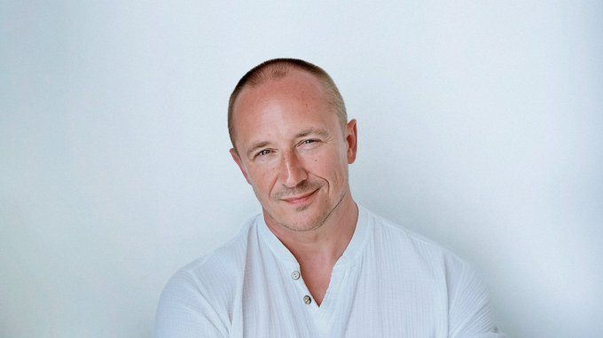 A portrait of a smiling man in a white henley shirt against a light background, appearing to be a professional headshot likely of a team member or founder associated with the Sleepagotchi project being discussed in the post.