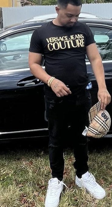 A man poses in front of a black car wearing a black "VERSACE JEANS COUTURE" t-shirt, distressed black jeans, and white sneakers, holding a Burberry crossbody bag. The image complements the post's giveaway announcement, showcasing a stylish look to engage followers.