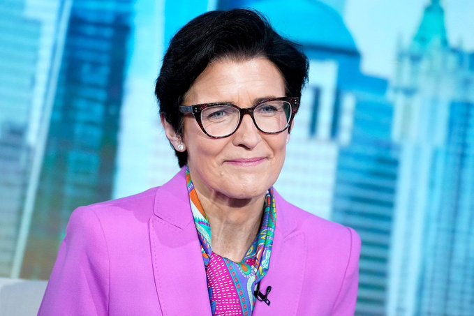 A professional portrait of a woman with short dark hair wearing black-rimmed glasses, a bright pink blazer, and a colorful patterned scarf, seated in front of a backdrop showing blue-toned skyscrapers.