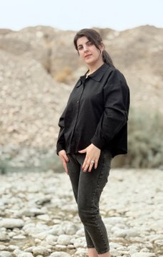 A woman with dark hair standing outdoors in a rocky landscape, wearing a black cropped jacket and dark jeans.