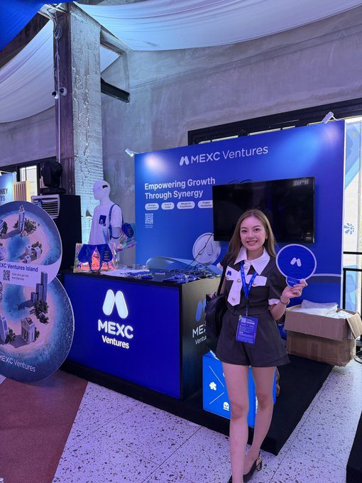 A woman in a black outfit stands at the MEXC Ventures 'Empowering Growth Through Synergy' booth, holding a branded fan at what appears to be a crypto industry event.