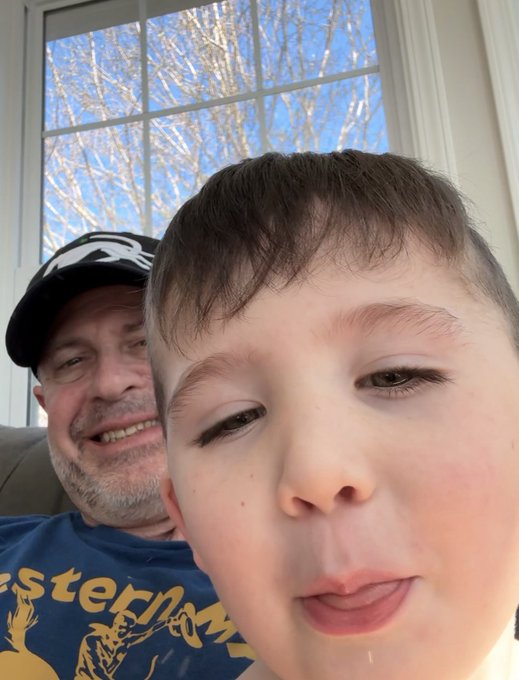 A selfie photo of a man wearing a black cap and a boy, taken indoors with a window showing bare trees and blue sky in the background.