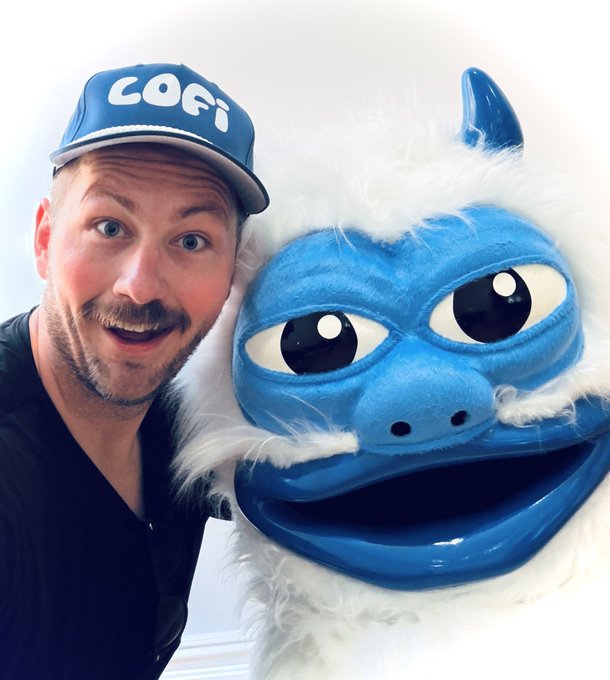 A man wearing a blue 'COFI' baseball cap posing with an excited expression next to a large blue and white furry mascot puppet.