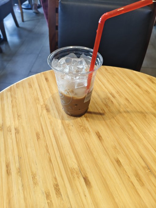 An iced coffee in a clear plastic cup with a red straw sits on a round bamboo table in what appears to be a cafe.