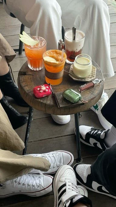 An overhead view of a small round wooden café table holding several beverages including an orange drink, a latte, and a small white cup, surrounded by the legs and feet of patrons in white Converse sneakers and black boots.
