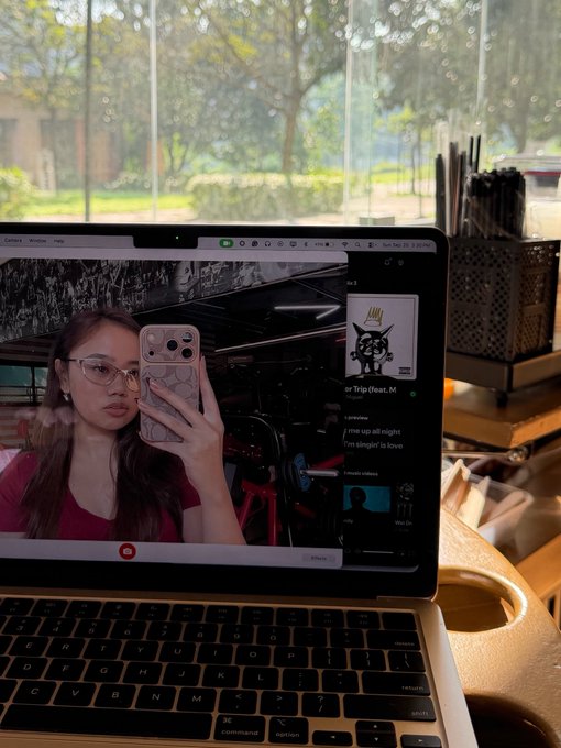 A MacBook laptop screen shows a video call where a woman with glasses is taking a mirror selfie with her iPhone, with a Spotify window displaying a song called 'me up all night' from the album 'im sleppin' in love'.