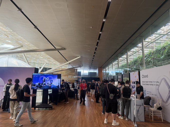 A wide indoor shot of a tech or crypto conference shows attendees walking through a modern venue with high ceilings; visible booth signage includes 'Qbond' reading 'Nebs make money from you. Davut makes money for you.'
