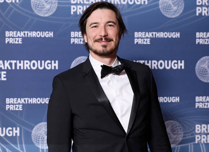 A man in a black tuxedo with a bow tie posing on the red carpet at the Breakthrough Prize event.