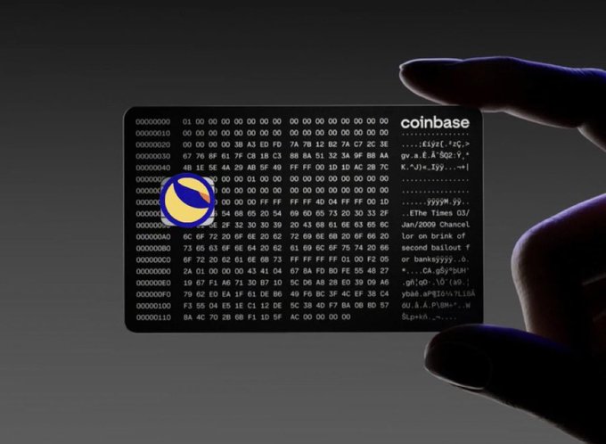A hand reaches toward a Coinbase debit card against a dark background; the card features the Coinbase logo, a grid of hexadecimal code, and the visible text '.EThe Times 03/Jan/2009 Chanclor on brink of second bailout f or banks', referencing the Bitcoin genesis block.