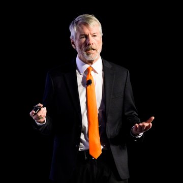 A man with gray hair and a beard, wearing a dark suit with an orange tie, speaks while gesturing with a presentation clicker against a black background. The image accompanies a statement from Michael Saylor about investing in Bitcoin as automation drives the value of traditional goods to zero.