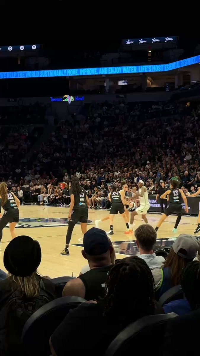 The image shows a basketball game taking place, with players wearing black uniforms on the court. The crowd of spectators can be seen watching the game from their seats. A man sitting in one of the chairs has his back turned towards the camera, while another person is seated closer to the front row and appears to be actively engaged in the game by looking at the players.