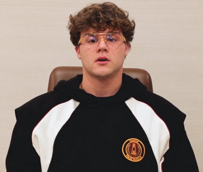 A young man with curly brown hair wears clear safety glasses and a black and white hoodie featuring an AC Milan logo, sitting in a brown leather chair.