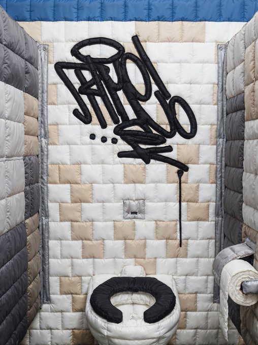 An inflatable/puffy art installation of a bathroom featuring a toilet and toilet paper roll, with black graffiti-style text reading 'SNOO BOO A...' on the back wall.