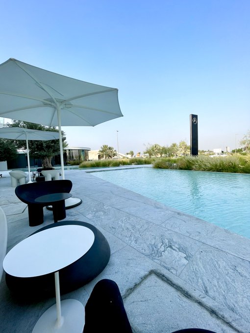 An outdoor patio scene featuring a swimming pool, white umbrellas, modern black and white lounge furniture, and landscaped grounds under a clear blue sky.