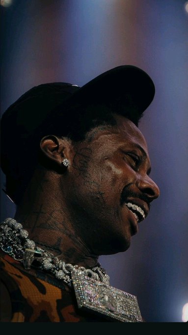 Profile photo of a smiling man wearing a black cap, diamond earrings, and diamond chains, with visible tattoos on his face and neck.