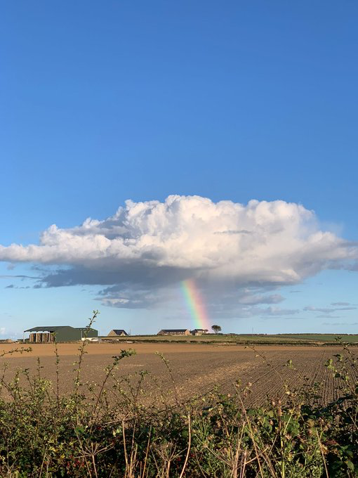 A scenic landscape featuring a rainbow descending from a large white cloud onto flat farmland, with farm buildings visible in the distance under a blue sky.