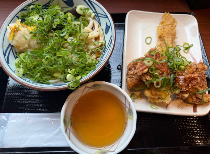The image shows a plate of food with noodles, chicken, and vegetables. There are two bowls - one containing soup or broth and another bowl filled with fried food. The dish appears to be a combination of Chinese cuisine, featuring both savory and flavorful elements.
