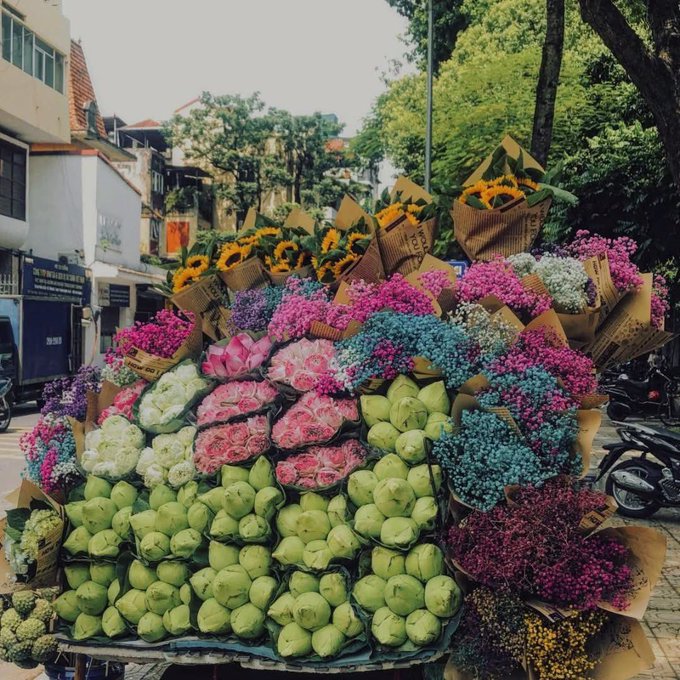 A vibrant Vietnamese street flower vendor display featuring bouquets of sunflowers, pink roses, purple blooms, and green lotus buds wrapped in paper, set against a backdrop of old shophouses.