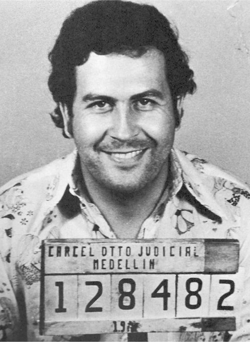 A black-and-white mugshot shows a smiling man with a mustache holding a placard reading 'CARCEL OTTO JUDICIAL MEDELLIN 128482,' identifying him as Pablo Escobar.