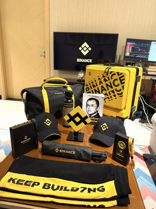 A collection of Binance branded merchandise, including a yellow suitcase, caps, duffel bag, tumbler, and a "KEEP BUILD7NG" banner, is displayed in front of a TV showing the Binance logo, marking the #binanceturns9 celebration with @BinanceAngels as part of a "Treasure Hunt" event.