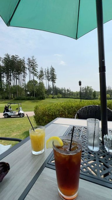 An outdoor patio scene shows three drinks on a metal table beneath a teal umbrella, with a view of a golf course, trees, and parked golf carts in the background.