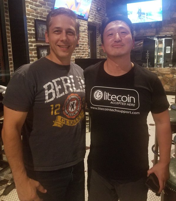 A photograph of two smiling men posing together at night; one wears a gray 'BERLIN' t-shirt while the other wears a black t-shirt promoting 'litecoin ACCEPTED HERE' with the URL 'www.litecointechsupport.com'.
