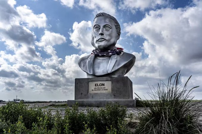 A silver bust statue of a man wearing an American flag bandana, mounted on a concrete pedestal labeled 'ELON, aka MemeLord, Pay Elon Musk no taxes,' set against a cloudy sky.