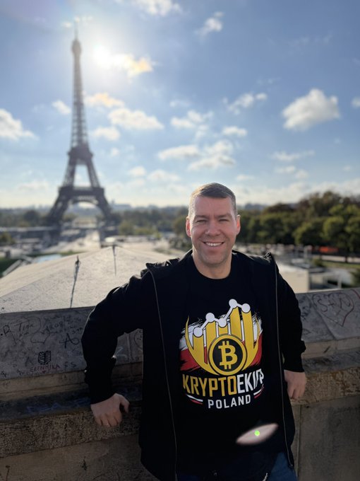 A smiling man wearing a black hoodie with a 'KRYPTOEKI.PL POLAND' Bitcoin-themed design, posing in front of the Eiffel Tower in Paris on a sunny day.