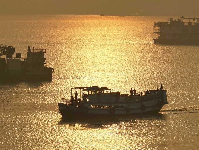 A golden silhouette photograph of two boats on shimmering water during sunset, with passengers visible on the closer vessel and what appears to be a floating structure on the right.