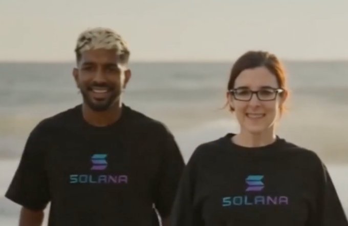 A smiling man and woman stand on a beach wearing black t-shirts featuring the 'SOLANA' logo.