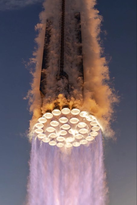 A SpaceX Starship rocket lifts off, its massive engines glowing bright orange against a dark blue sky as exhaust and steam billow out beneath it.