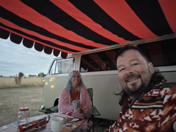Two people smiling under a red and black striped awning next to a vintage van, with one person wearing a pink cardigan and the other in a patterned jacket.