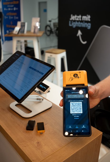 A hand holds a smartphone displaying the BitBoxApp with a Bitcoin Lightning payment QR code next to a point-of-sale tablet, illustrating the Lightning-powered Bitcoin payment setup tested at the Blocktrainer event, with a "Jetzt mit Lightning" (Now with Lightning) sign visible in the background.