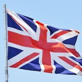 The Union Jack flag of the United Kingdom is shown waving against a light sky.