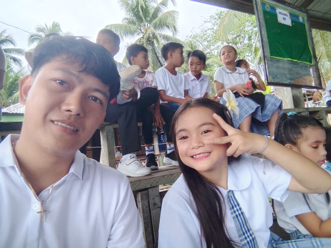A selfie of a smiling young man and woman making a peace sign, taken outdoors with other people in school uniforms sitting on benches in the background near tropical trees.