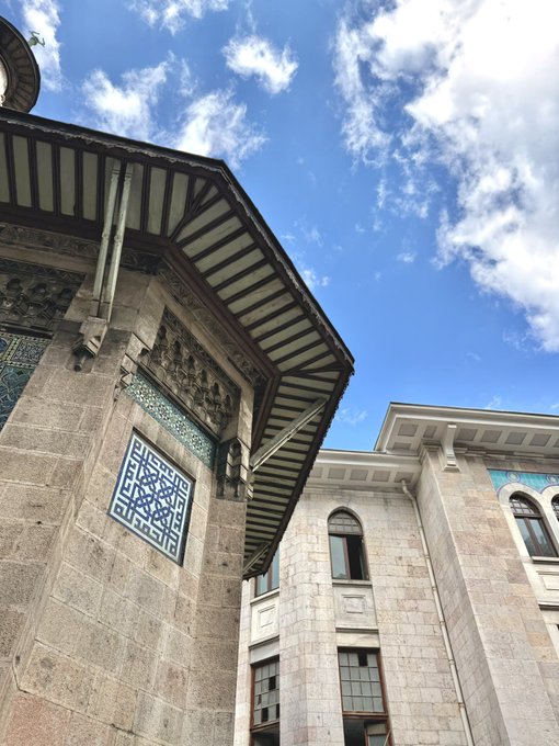 A low-angle architectural photograph showing traditional Ottoman-style buildings with detailed tilework and wooden eaves set against a bright blue sky with scattered clouds.