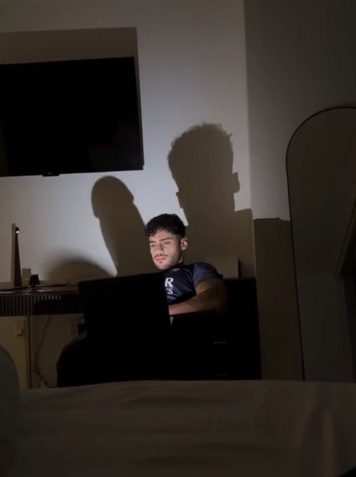A young man sits in a dimly lit room with a laptop, his face illuminated by the screen light casting dramatic shadows on the wall behind him.