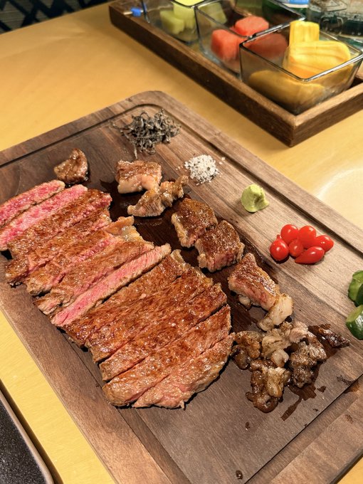 A beautifully sliced medium-rare steak is presented on a wooden cutting board, garnished with wasabi, cherry tomatoes, salt, and herbs, accompanied by a tray of fresh fruit in the background. The image perfectly captures the "币安上牛来，买了牛来的吃牛来" sentiment—showcasing an indulgent, premium beef meal enjoyed after the post.