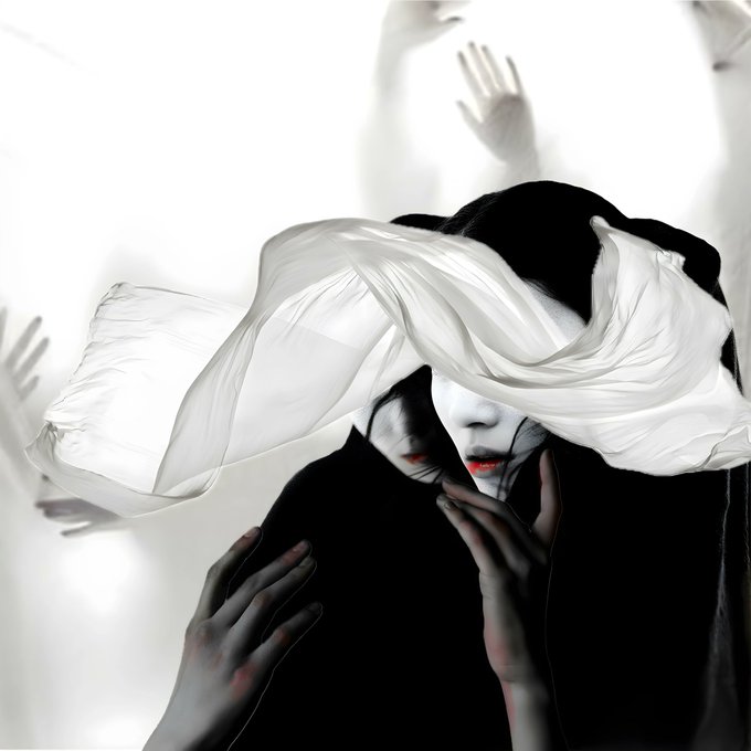 An artistic black-and-white photograph of a person wearing a white robe covering their head, with their face partially visible showing red lips, and hands reaching toward them in the background.