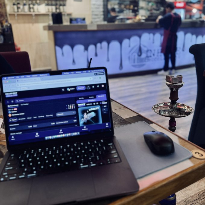 A MacBook laptop displaying a Spotify playlist alongside a wireless mouse and a decorative hookah, set on a table in what appears to be a café or lounge.