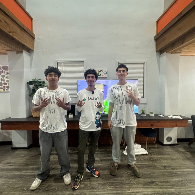 Three young men pose making hand gestures while wearing matching white t-shirts with hand-drawn designs, standing in a modern office/living space.