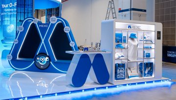 A blue and white MEXCO exhibition stand at a trade show, showcasing products on shelves and featuring a triangular logo design.