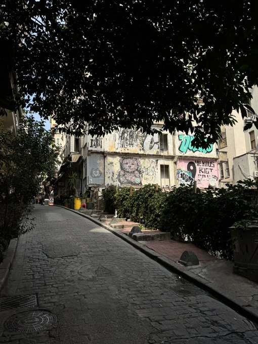 A narrow cobblestone street lined with trees, leading to a wall covered in colorful graffiti and street art under a partially shadowed view.