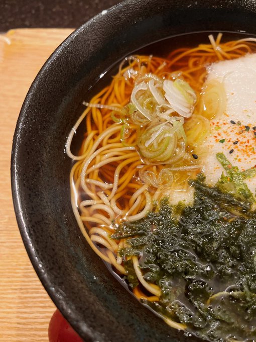 A close-up of a bowl of ramen with thin noodles, scallions, a poached egg, seaweed, and a sprinkle of chili pepper in a dark broth.