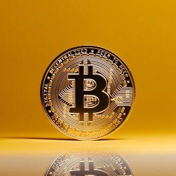 A gold Bitcoin token stands upright against a yellow background, displaying the text "BITCOIN DIGITAL * DECENTRALIZED * PEER TO PEER" around its edge. The image visually represents the article's discussion of Bitcoin's price floor and the cryptocurrency's struggle to break above $80K without a shift in monetary policy.