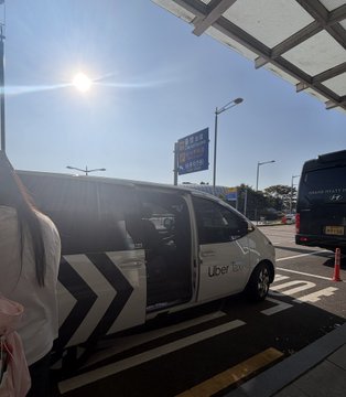Photograph of an Uber Taxi van parked at what appears to be an airport taxi stand on a sunny day, with a person's hair visible in the foreground.