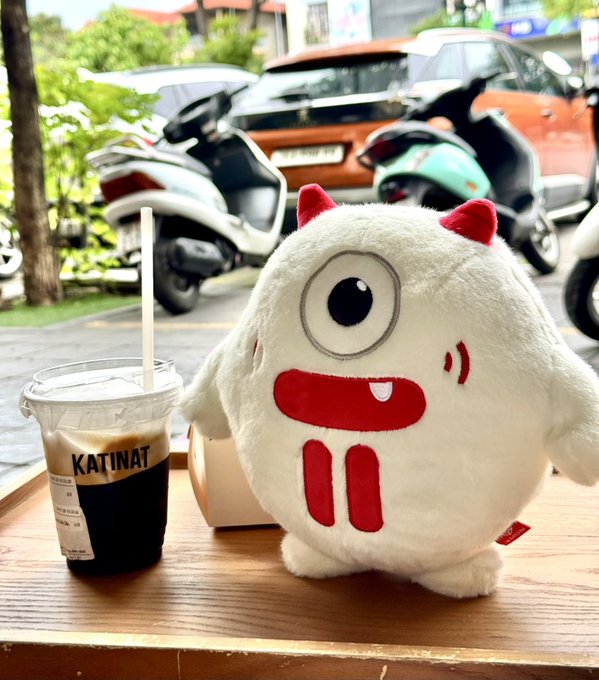 A white plush monster character with one large eye and red horns sits on a wooden table next to a plastic cup of dark drink with a 'KATINAT' label, with motorcycles parked outside the window.